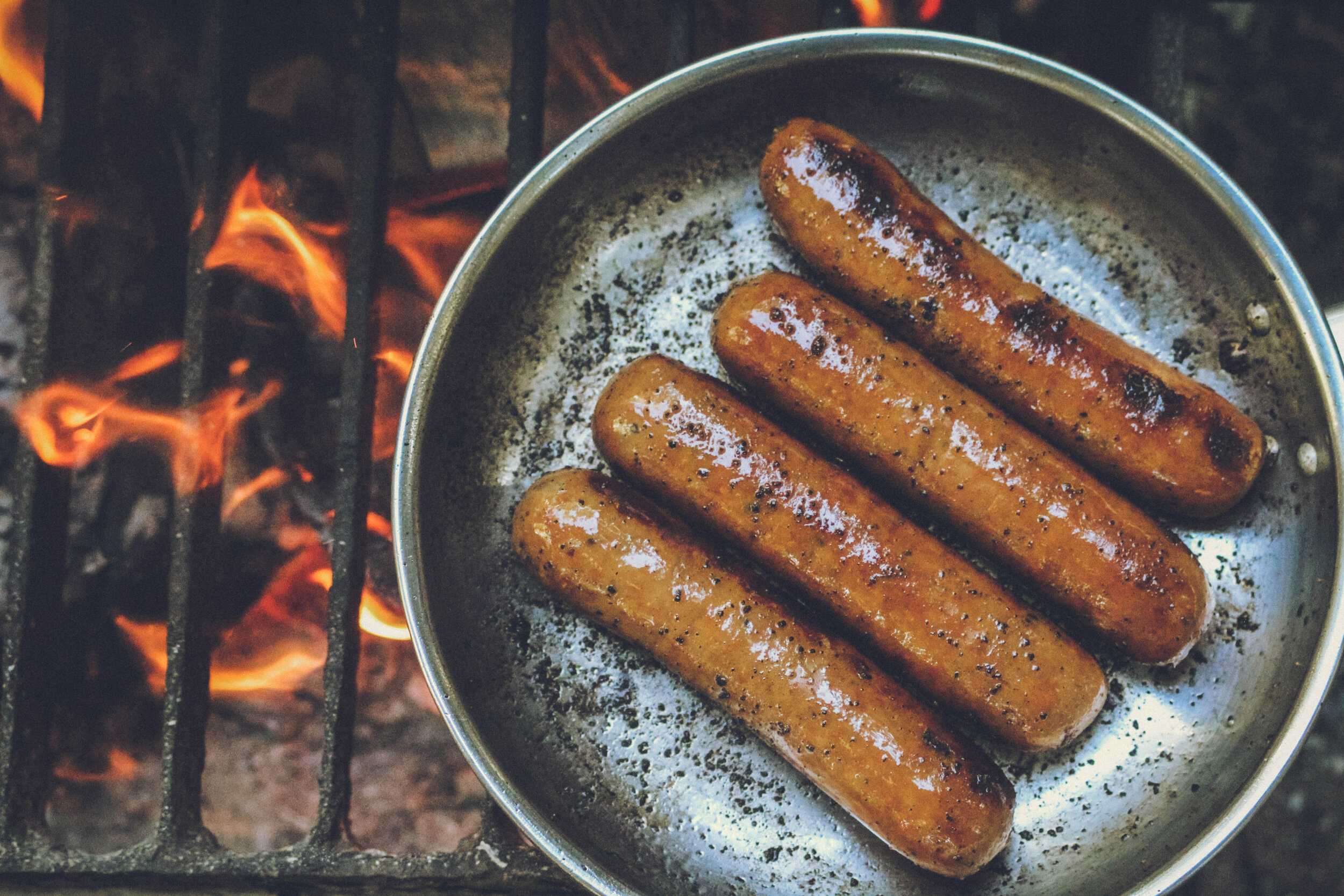 Grilled sausages cooking in a metal pan over an open flame, showcasing a delicious caramelized exterior and smoky flavor. Perfect for outdoor barbecues and summer gatherings.